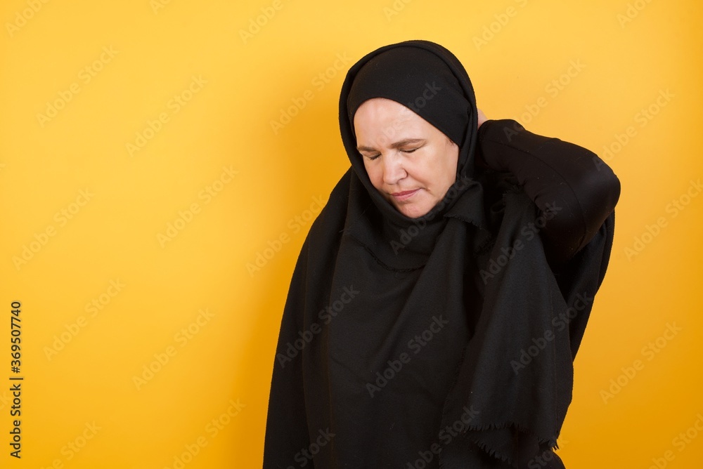Beautiful middle aged muslim woman wearing black hijab over yellow background Suffering of neck ache injury, touching neck with hand, muscular pain Beautiful middle aged muslim woman wearing black hijab over yellow background Suffering of neck ache injury, touching neck with hand, muscular pain