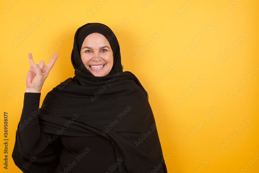 Middle aged muslim woman wearing black hijab over yellow background doing a rock gesture and smiling to the camera. Ready to go to her favorite band concert. Middle aged muslim woman wearing black hijab over yellow background doing a rock gesture and smiling to the camera. Ready to go to her favorite band concert.