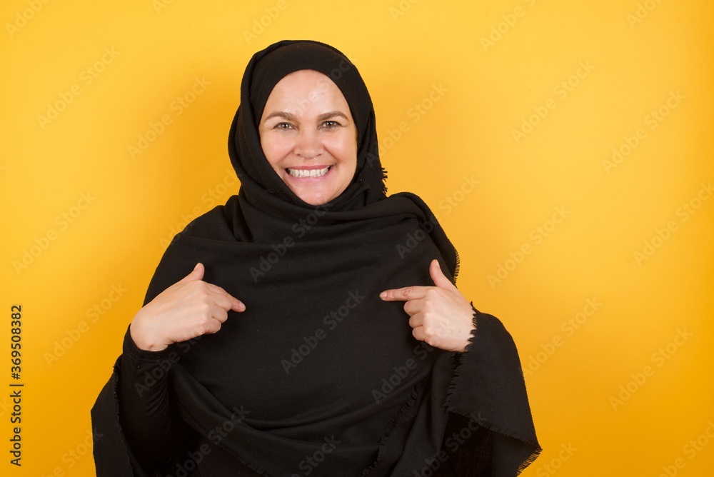 Horizontal shot of cheerful Middle aged muslim woman wearing black hijab over yellow background points at his body, being in good mood after going shopping and making successful purchases Horizontal shot of cheerful Middle aged muslim woman wearing black hijab over yellow background points at his body, being in good mood after going shopping and making successful purchases