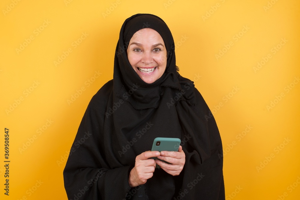 Copyspace photo of cheerful nice Middle aged muslim woman wearing black hijab over yellow background with collar holding phone in his hands while Copyspace photo of cheerful nice Middle aged muslim woman wearing black hijab over yellow background with collar holding phone in his hands while
