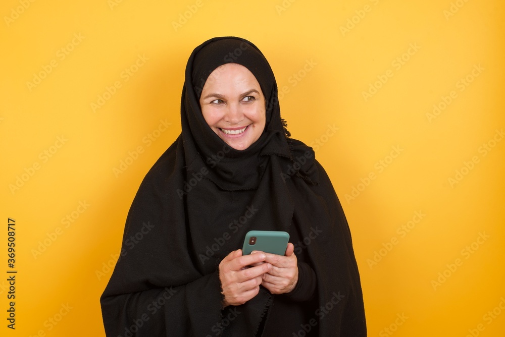 Middle aged muslim woman wearing black hijab over yellow background telephone hands read good youth news look empty space advert Middle aged muslim woman wearing black hijab over yellow background telephone hands read good youth news look empty space advert
