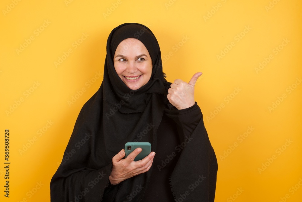 Middle aged muslim woman wearing black hijab over yellow background using and texting with smartphone pointing and showing with thumb up to the side with happy face smiling Middle aged muslim woman wearing black hijab over yellow background using and texting with smartphone pointing and showing with thumb up to the side with happy face smiling