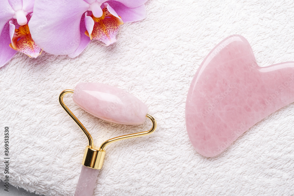 Rose Quartz jade roller and Gua Sha massager on towel on stone background. Close-up. Massage tool for facial skin care, SPA beauty treatment concept Rose Quartz jade roller and Gua Sha massager on towel on stone background. Close-up. Massage tool for facial skin care, SPA beauty treatment concept