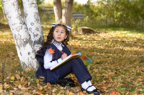 A schoolgirl with a book sits under a birch tree in an autumn forest. Elementary school student in the school park. A girl in a blue school uniform in an autumn park looks at the sky
