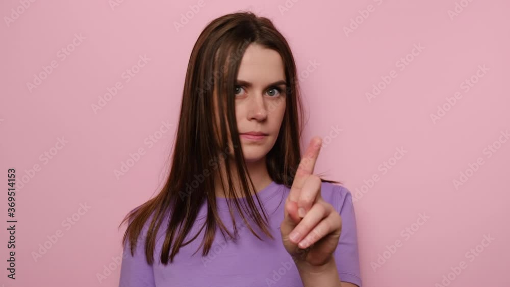 Close up portrait of young woman warning with admonishing finger gesture, saying no, giving advice to avoid danger, dressed in purple t-shirt, disapproval sign, isolated on pink studio background