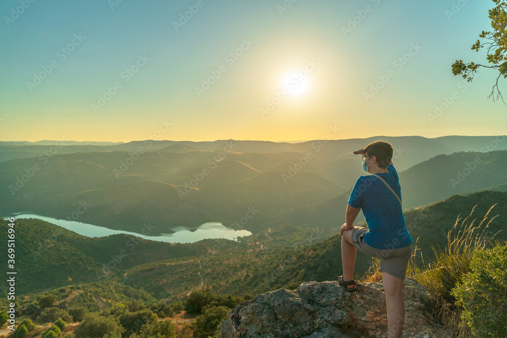 Fototapeta premium Young hiker with surgical mask watches the sunset from the top of the valley relaxing with the river Duero and the mountains, Vilvestre, Spain