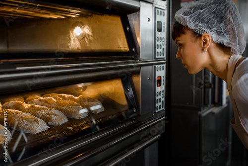 Young caucasian woman baker is looking at the bread baker process in an electric oven at a baking manufacturing factory.