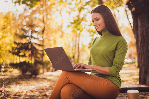 Photo of pretty cute lady smiling sit bench park hold laptop drink coffee remotely work project programming app communicate chatting client wear green turtleneck orange pants outdoors