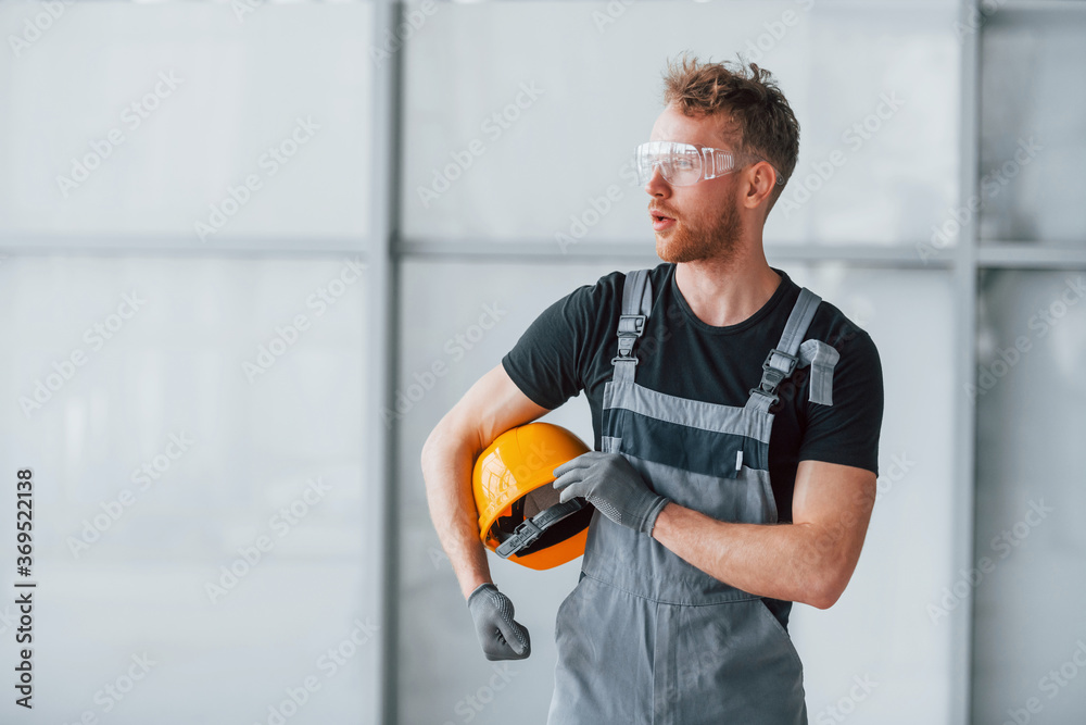 Portrait of man in grey uniform andwith orange hard hat in hands that standing indoors in modern big office at daytime Portrait of man in grey uniform andwith orange hard hat in hands that standing indoors in modern big office at daytime