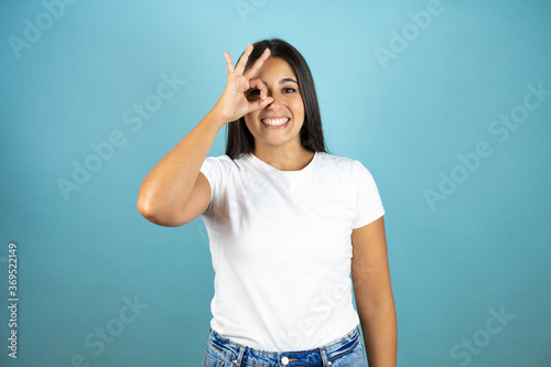 Young beautiful woman standing over isolated blue background doing ok gesture shocked with smiling face, eye looking through fingers © Irene