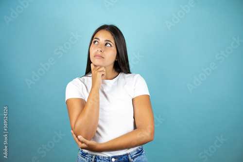 Young beautiful woman standing over isolated blue background looking side confident, thinking with crossed arms and hand raised on chin