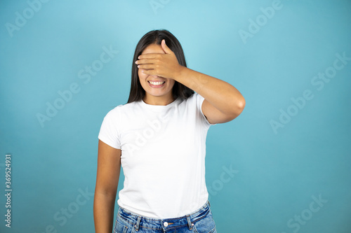 Young beautiful woman standing over blue isolated background smiling and covering her eyes with her hand