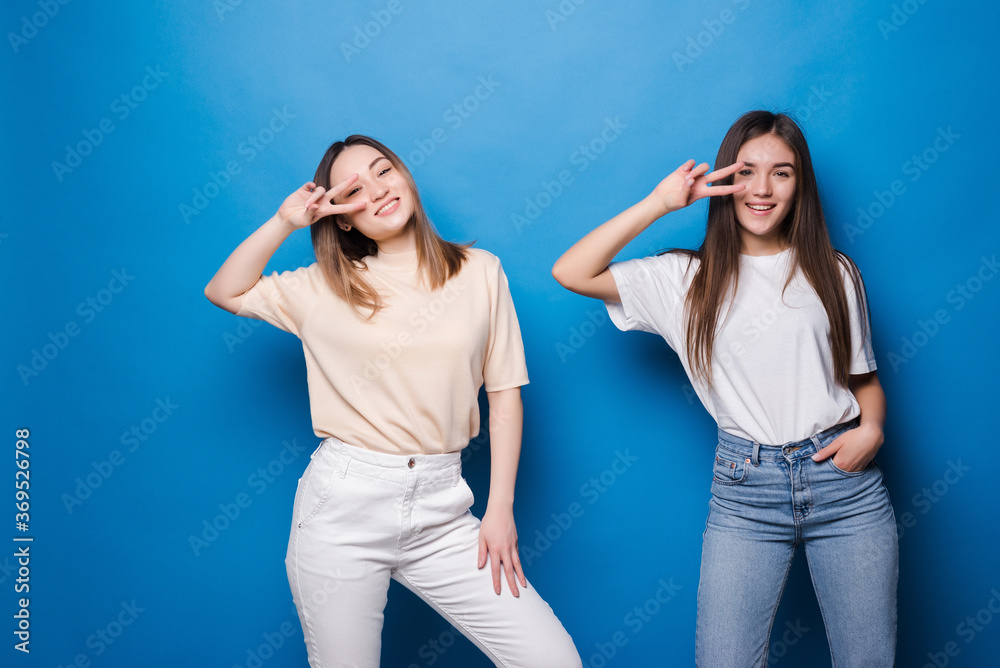 Picture of two playful girls standing together and showing peace gestures over blue background