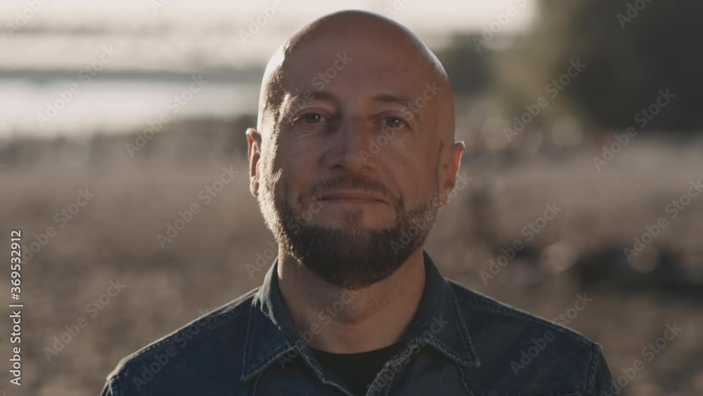 Bald hipster in his mid forties with a stylish beard is standing on the beach during a beautiful sunset. He is smiling gently. Bald hipster in his mid forties with a stylish beard is standing on the beach during a beautiful sunset. He is smiling gently.