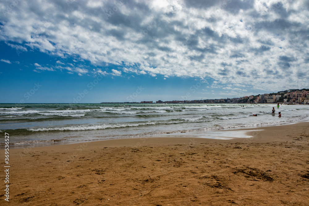 Fototapeta premium Nettuno, Lazio, Rome, Italy - Some people bathe, play and relax in the water, in the Roman sea. Reflections of light on the water. Cloudy blue sky at sunset in summer. Waves of the sea slightly choppy