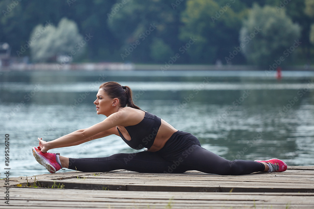 Attractive girl in sportswear does gymnastic exercises on a wooden pier in a city park.