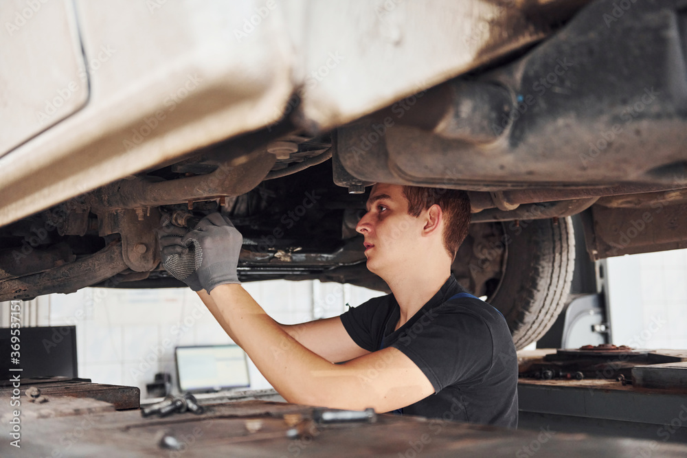 Professional repairman in black shirt works under broken automobile Professional repairman in black shirt works under broken automobile