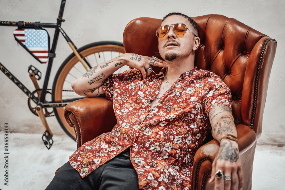 Very expressive and stylish guy in hawaiian shirt and sunglasses relaxing on a retro sofa looks like freakly gangster | Adobe Stock