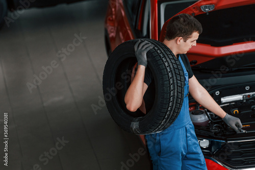 Worker in black and blue uniform holds car wheel and walks with in near red automobile