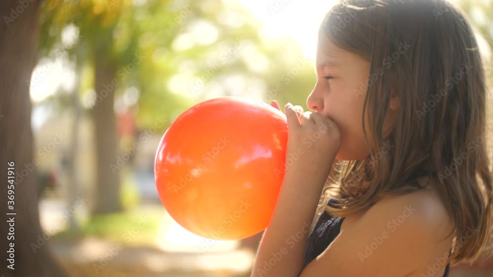 Cute 7 year old girl child blowing up inflatable orange balloon in summer street. Shallow DOF, slow motion.
