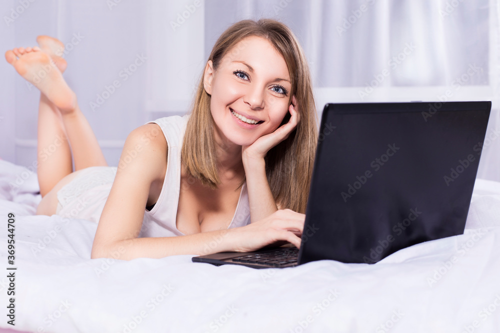 Beautiful blonde woman working on a laptop in her bed in pajamas, smiling Beautiful blonde woman working on a laptop in her bed in pajamas, smiling