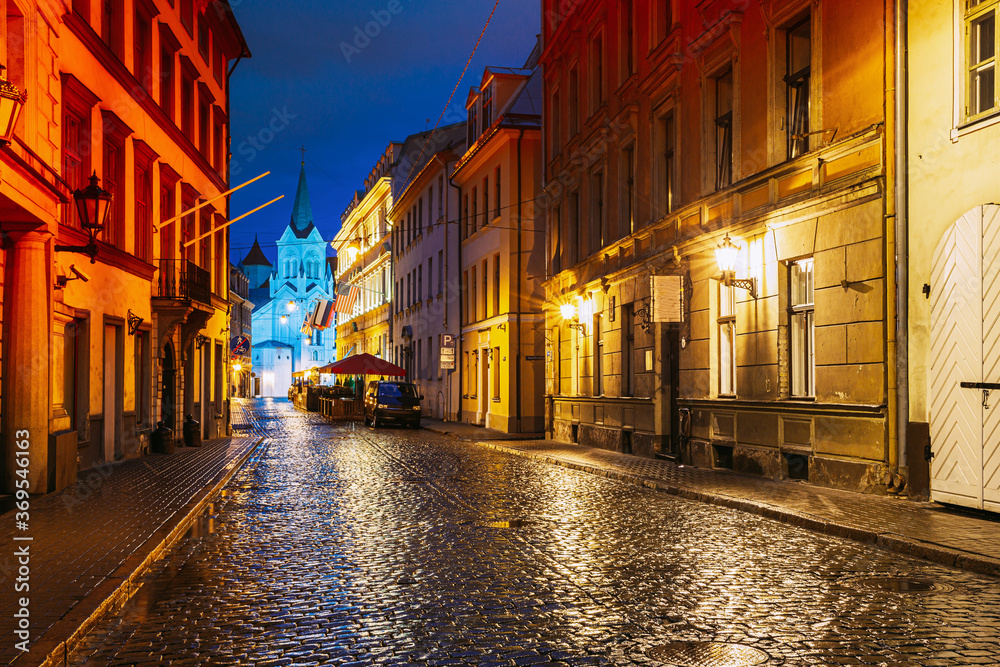 Riga, Latvia. Evening View Of Pils Street With Ancient Architecture In Bright Yellow Illumination Under Summer Blue Sky. Our Lady Of Sorrows Or Virgin Of Anguish Church In Distance