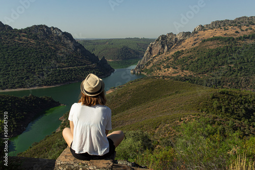 Attractive young female with a hat sitting on top of a rock in a viewpoint witnessing a river flowing between the mountains in Monfrague, Spain.
