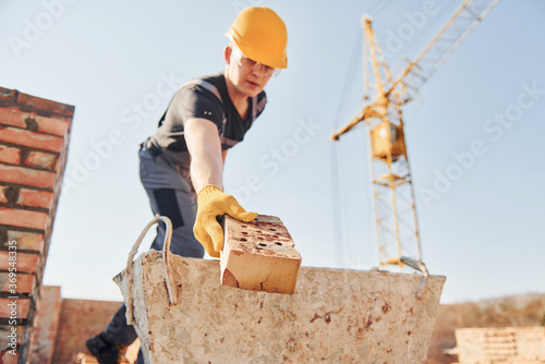Holding brick and using hammer. Construction worker in uniform and safety equipment have job on building