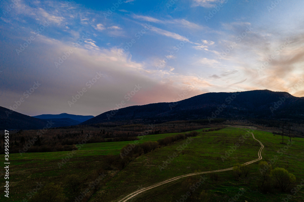 Carpathian meadow and beautiful sunset in the mountains - spring landscape, spruces on the hills, dark cloudy sky and bright sunlight, the village can be seen on the horizon.