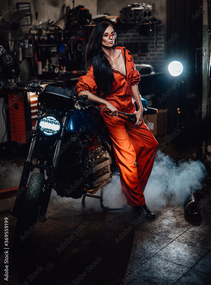 Sexy looking brunette in work overalls and protective googles leaning on a black motorcycle in a car service