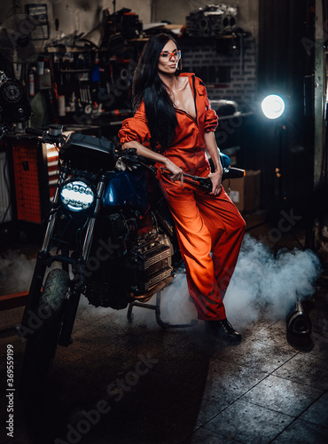 Sexy looking brunette in work overalls and protective googles leaning on a black motorcycle in a car service