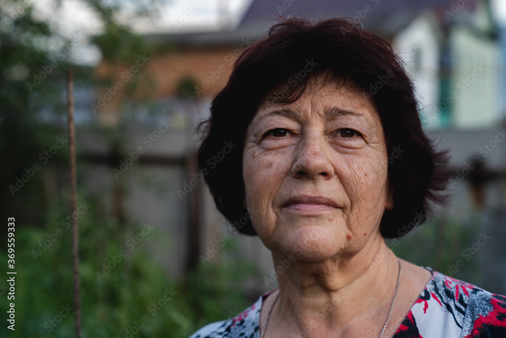 Senior woman portrait. She is outside on her back yard in a village. She seriously looking at the camera. Natural appearance. Horizontal photo Senior woman portrait. She is outside on her back yard in a village. She seriously looking at the camera. Natural appearance. Horizontal photo