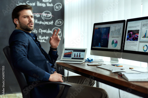 Young confident businessman in wireless earphones talking with client or partner, discussing sales report while working with statistical data in the modern office