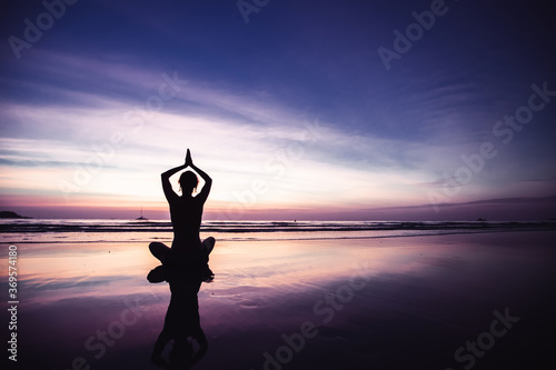 Silhouette of yoga woman in lotus position on the beach with reflection in water during twilight.