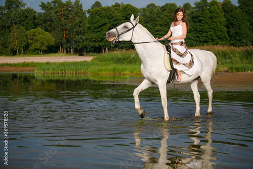 Young beautiful girl with white horse on the water at sunset or sunrise. Caucasian woman in dress in boho style on background at sea.Horseback rider on beach. Summertime scene © digitalien