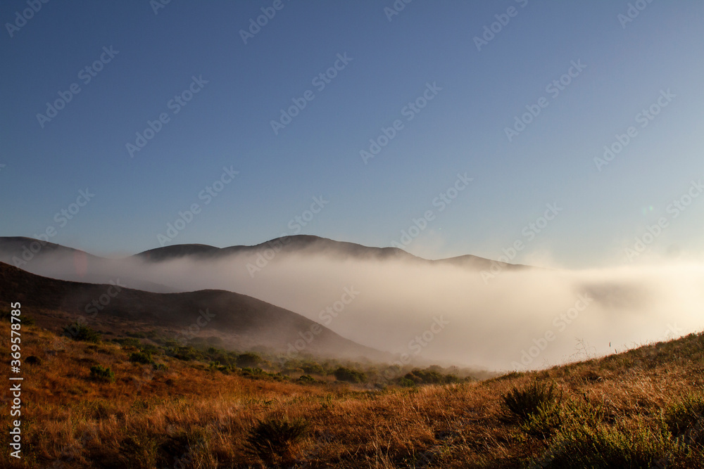 Fog rolls in at sunrise, desert plants are growing in the rocky, mountainous terrain.
