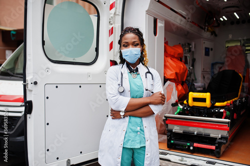 African american female paramedic in face protective medical mask standing in front of ambulance car. photo