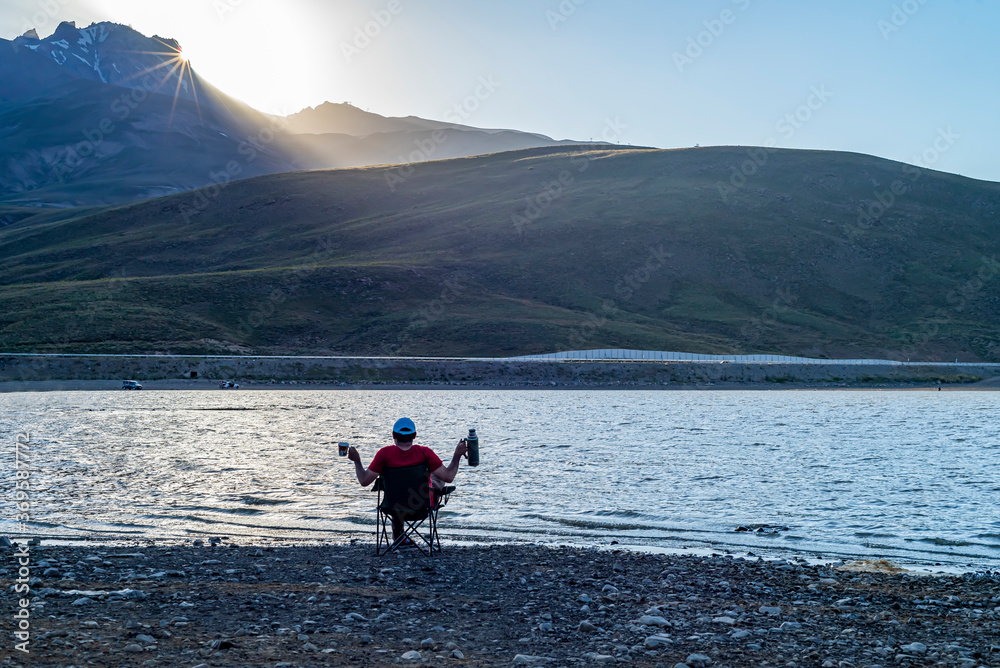 Camping area on a lake in mountainous terrain. A man with a camp chair and a tea thermos