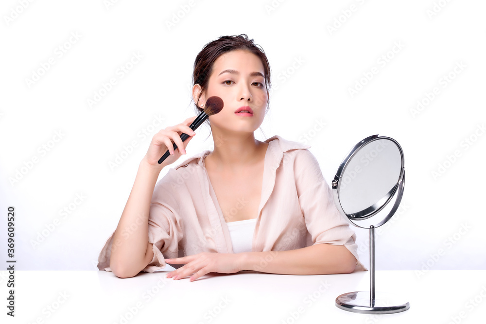 Portrait of beautiful charming young Asian woman brushing facial with cosmetic and sitting with white table, beauty of female face with skincare on white background, beauty cosmetic make up concept Portrait of beautiful charming young Asian woman brushing facial with cosmetic and sitting with white table, beauty of female face with skincare on white background, beauty cosmetic make up concept