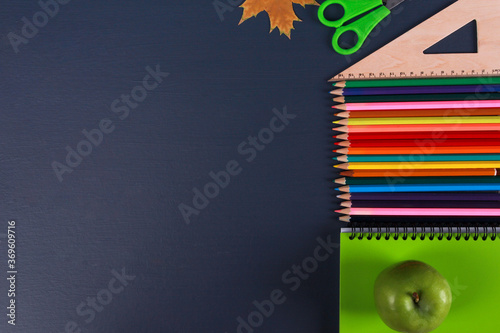 Green notebook, colorful pencils, fresh apple, ruler, autumn leaf on dark blue background. Top view, copy space, close-up. Back to school in September concept