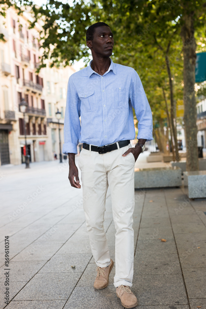 Full-length portrait of young african american man wearing light clothes walking along city street on summer day.