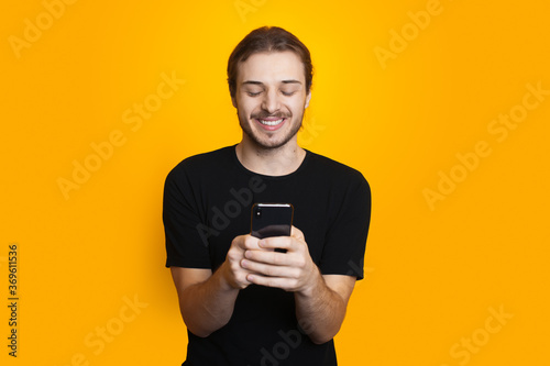 Bearded man with long hair is chatting on mobile in black clothes and smile on a yellow studio wall