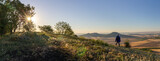 Young man with backpack standing on hill and looking to Czech central mountain valley at sunrise. Panoramic landscape
