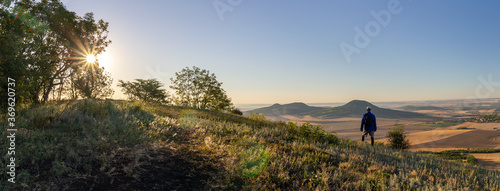 Young man with backpack standing on hill and looking to Czech central mountain valley at sunrise. Panoramic landscape