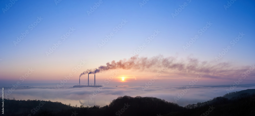 Panoramic view of three smoking stacks of thermal power station on the horizon taken from the hill, pipes are in evening fog on blue sky, copy space. Concept of ecology and environmental pollution.