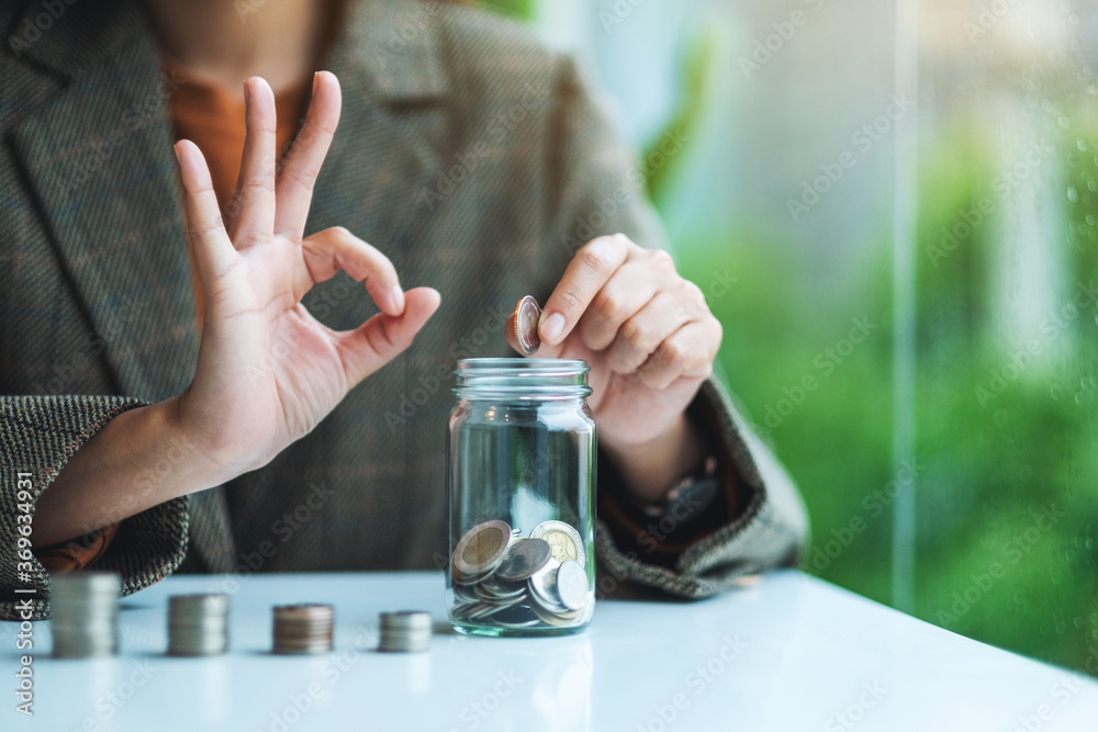 Closeup image of a businesswoman making ok hand sign while putting coins in a glass jar