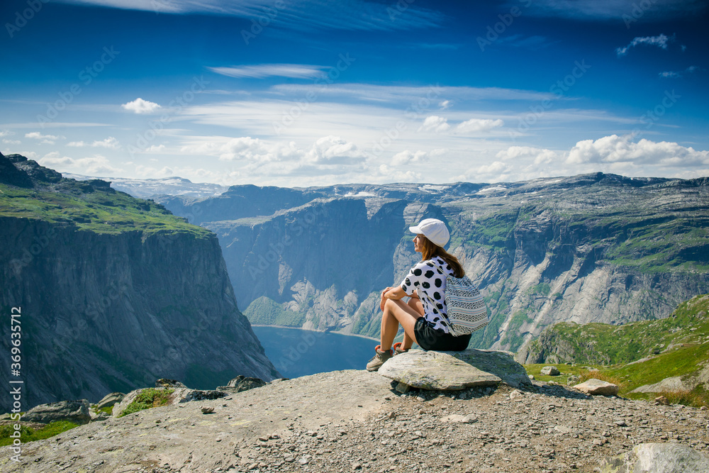 Natural landmark of Norway. Girl sitting on the edge of a rocky cliff. Extreme tourism. Girl with a backpack. Mountains and fjords of Norway. A tourist in a white cap on a cliff looks at the scenery