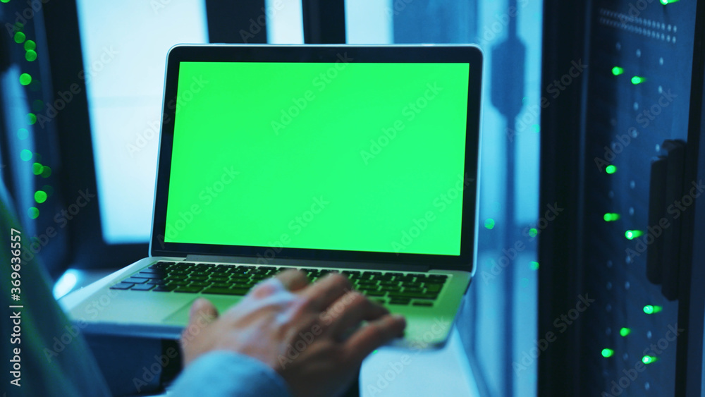 IT administrator using a mock-up laptop running diagnostics and web hosting with chrome key greenscreen device inside server cabinet of database. Close-up. Advanced data center. IT administrator using a mock-up laptop running diagnostics and web hosting with chrome key greenscreen device inside server cabinet of database. Close-up. Advanced data center.