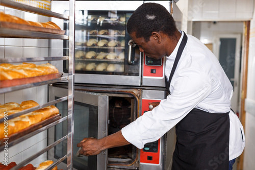 Portrait of African American baker controlling process of baking bread in professional oven at bakery