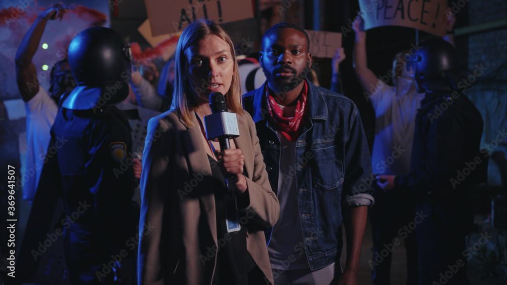 Beautiful serious woman journalist making a live speech on TV explaining the situation with night street demonstration rally. No racism. Black lives matter. Shocking news.